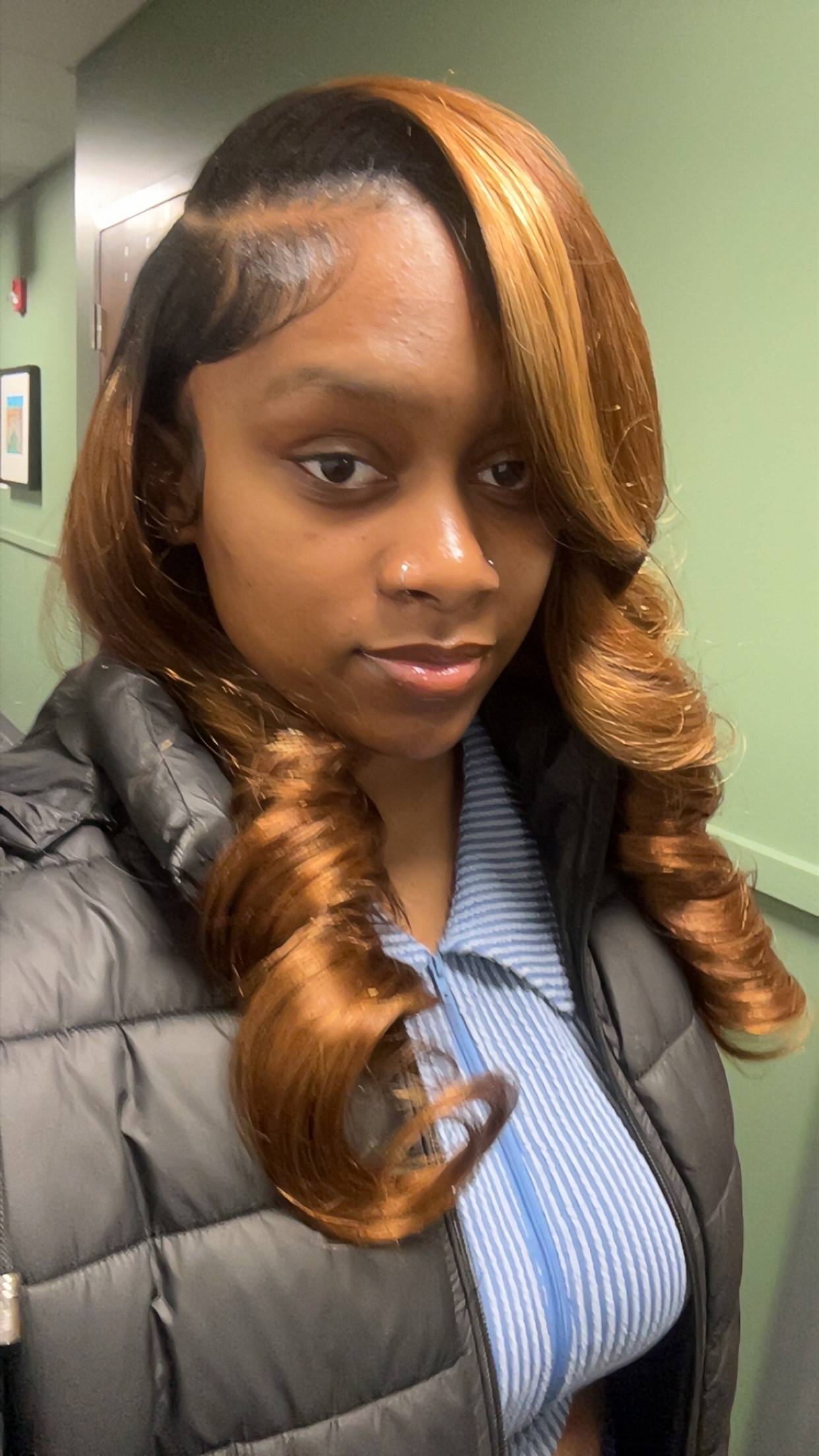Young woman smiling at the camera wearing a blue striped shirt and grey jacket with a green wall in the background